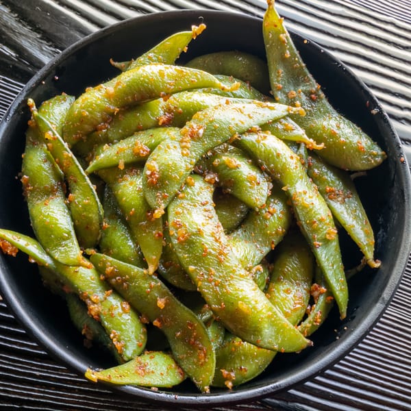 A black bowl filled with green edamame pods, seasoned with red spices, on a striped surface.