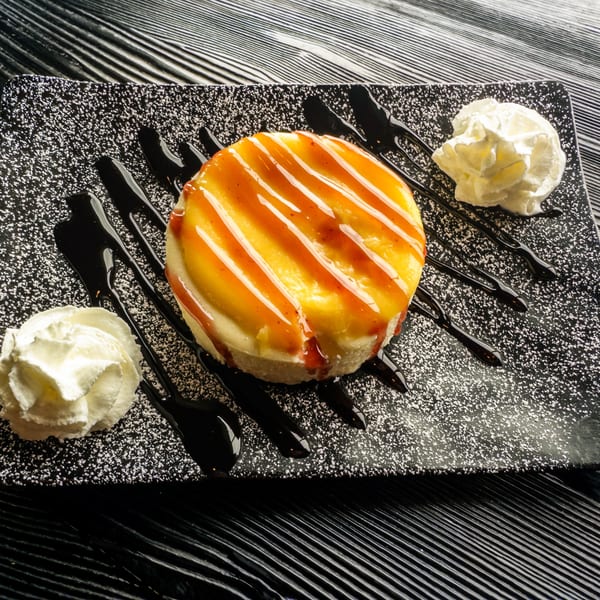 A cheesecake with orange and red drizzle, chocolate sauce, and whipped cream on a speckled black plate.