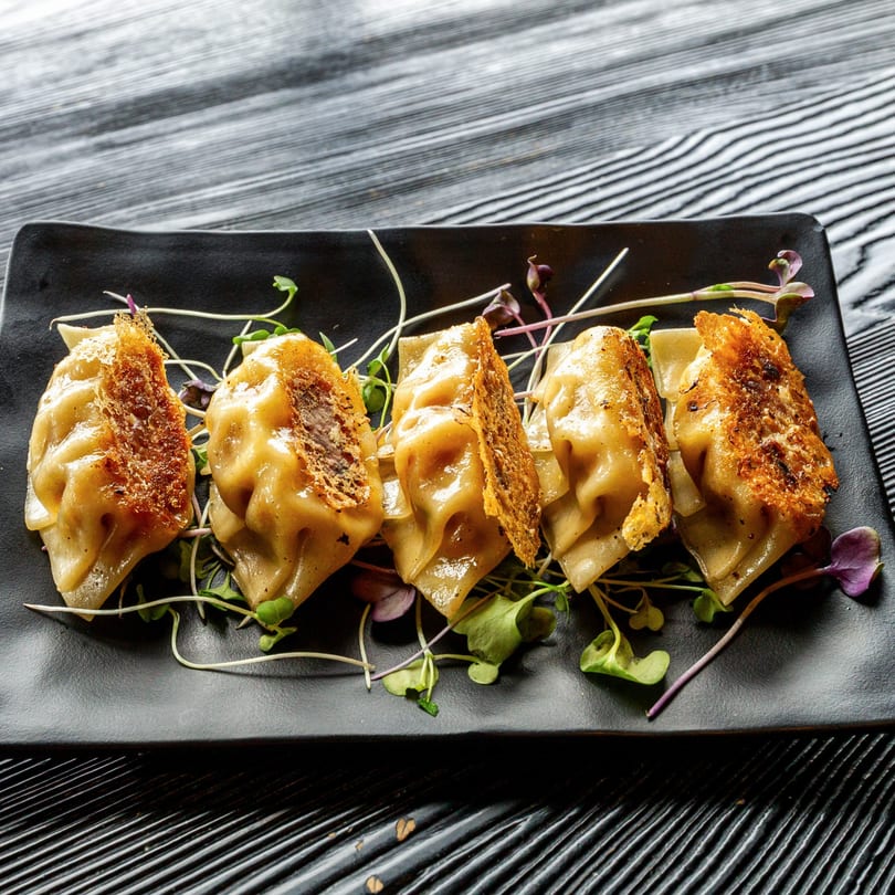 Five golden-brown Susukino Ramen Bar gyoza with crispy edges and microgreens on a black plate.