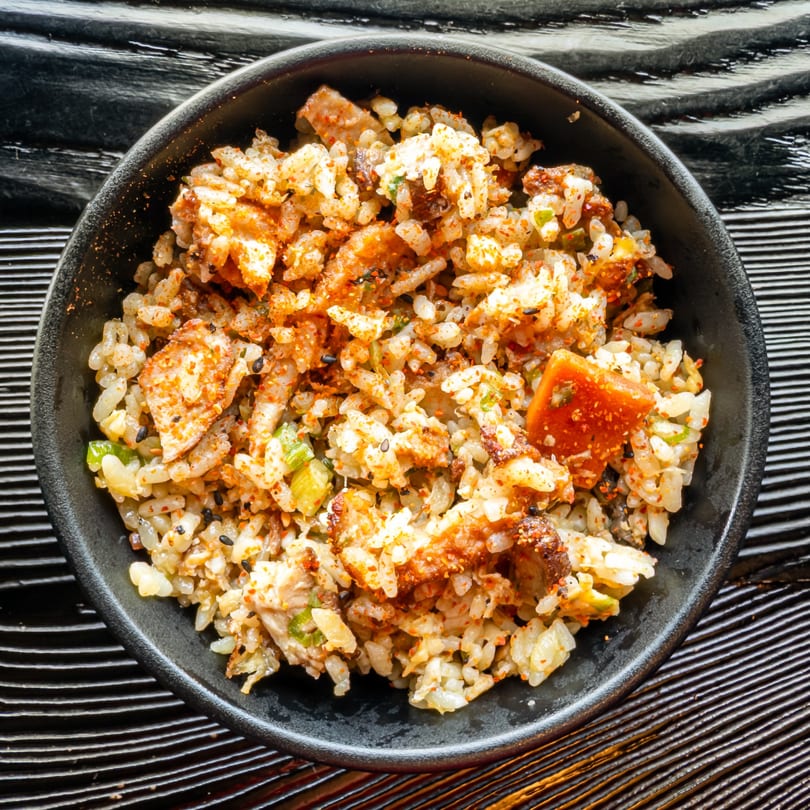 A close-up of Susukino Ramen Bar's fried rice with chicken, vegetables, and red spice in a black bowl.